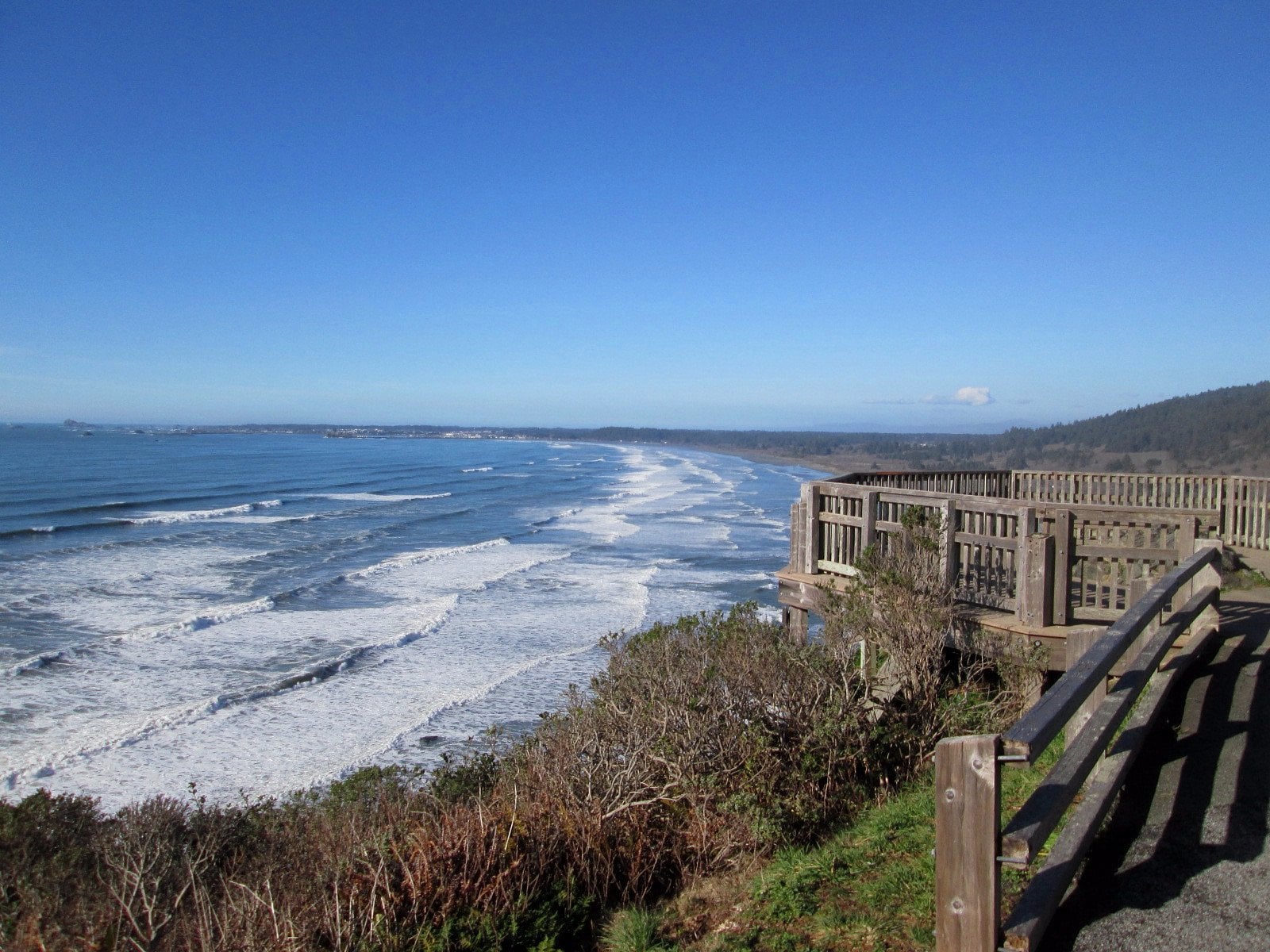 A wooden observation deck overlooks a scenic coastline with rolling waves, sandy beach, and distant headlands under a clear blue sky. Shrubs and greenery grow along the cliff edge.