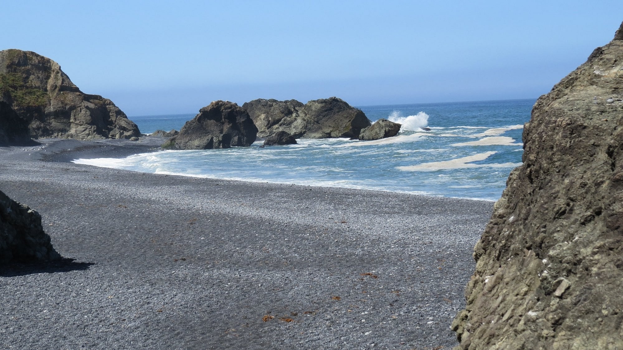 Rocky beach with dark pebbles, bordered by large cliffs and rocks. Ocean waves crash against the rocks under a clear blue sky. Sparse seaweed is scattered on the shore.