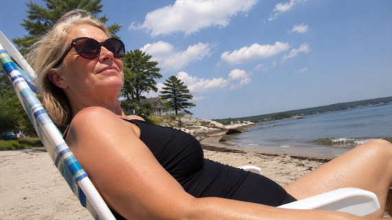 A woman in sunglasses and a black swimsuit relaxes on a beach chair, enjoying the sunny weather. Set against the backdrop of charming beach towns, the scene features the ocean, trees, rocky shoreline, and a blue sky with clouds.