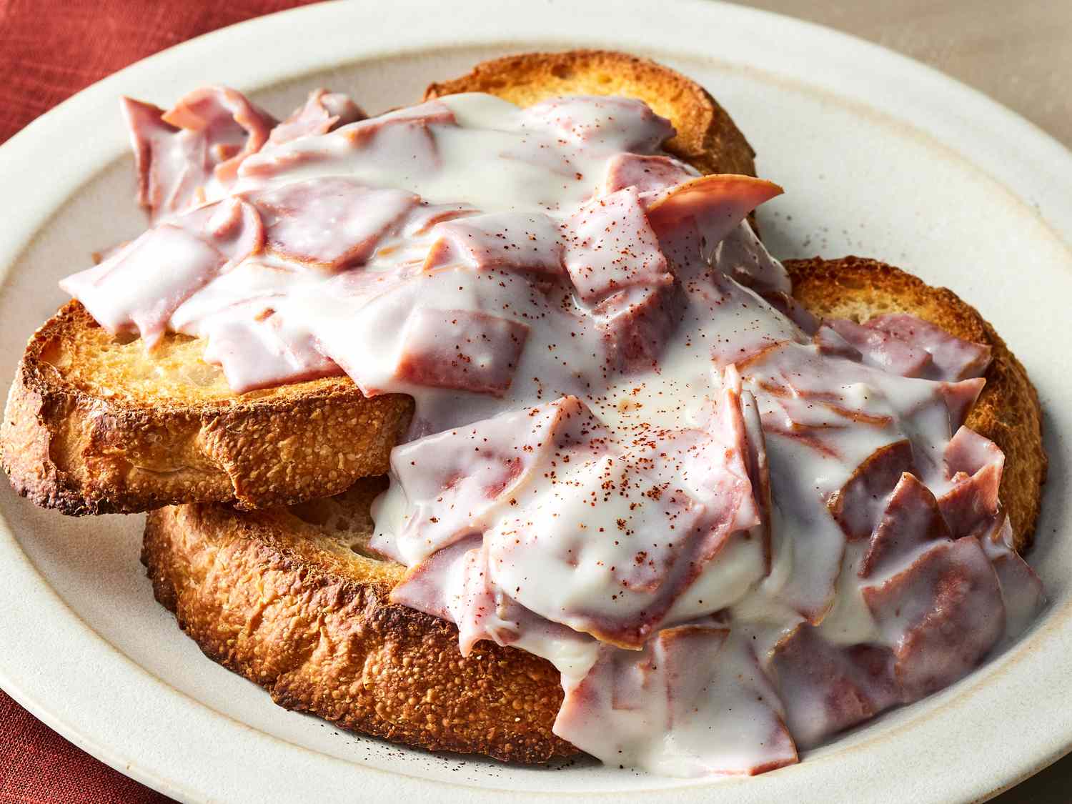 Two slices of toasted bread on a plate topped with chopped pieces of meat and creamy white sauce, lightly sprinkled with black pepper.