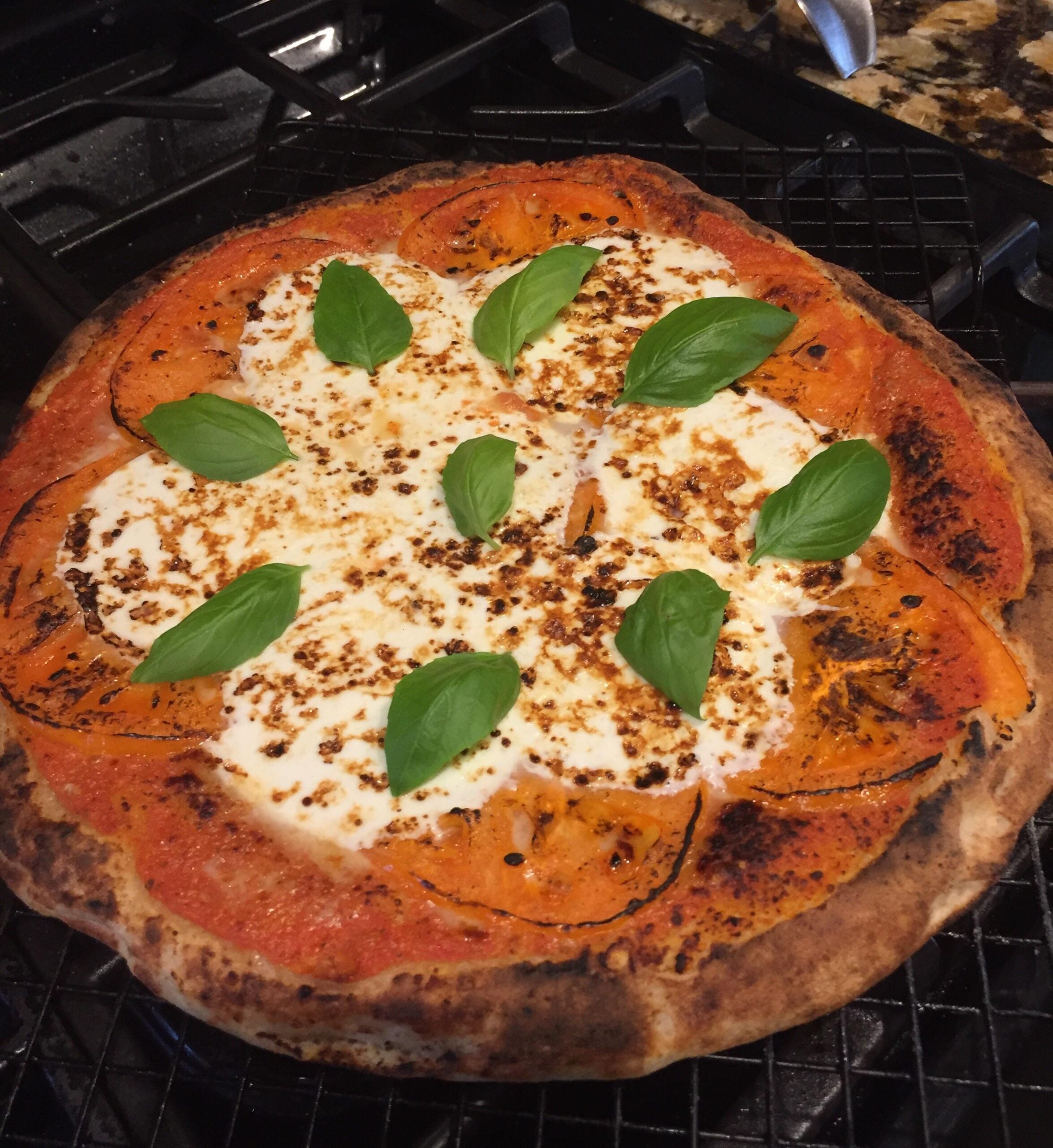 A freshly baked pizza with melted mozzarella, sliced tomatoes, and basil leaves on top, resting on a cooling rack near a stove.