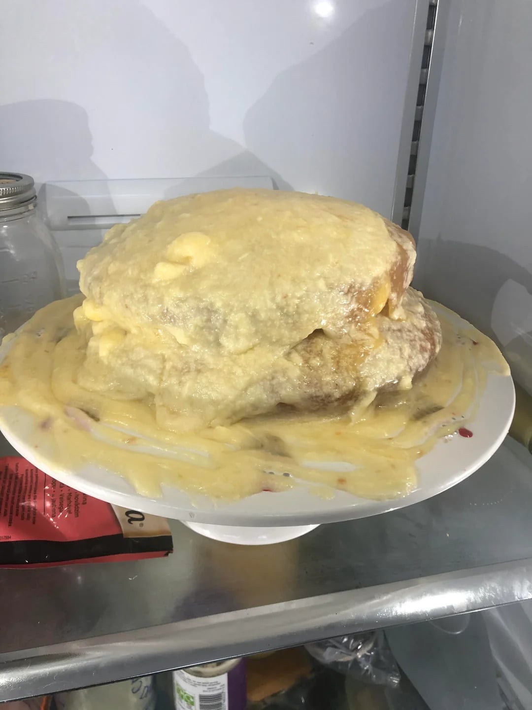 A cake on a white plate is covered in runny, dripping frosting, with the icing pooled on the plate and the cake sitting inside a refrigerator.