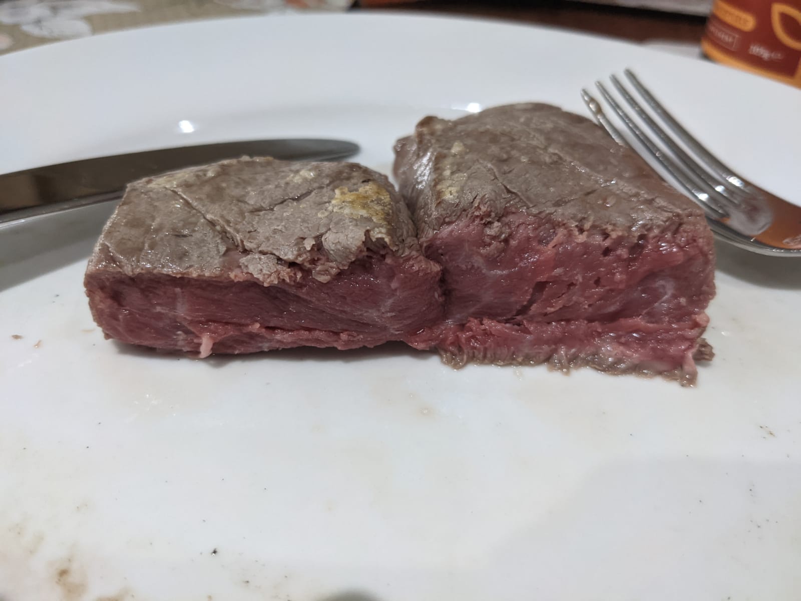 A close-up of a piece of beef steak cut in half on a white plate, showing a mostly raw, red interior. A fork and knife are placed next to the steak.