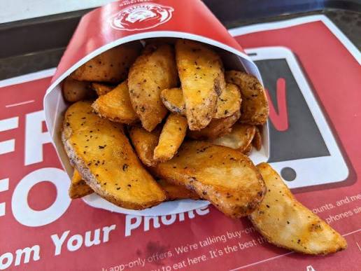 A container of seasoned potato wedges is tipped over, spilling crispy wedges onto a red tray liner with white text.