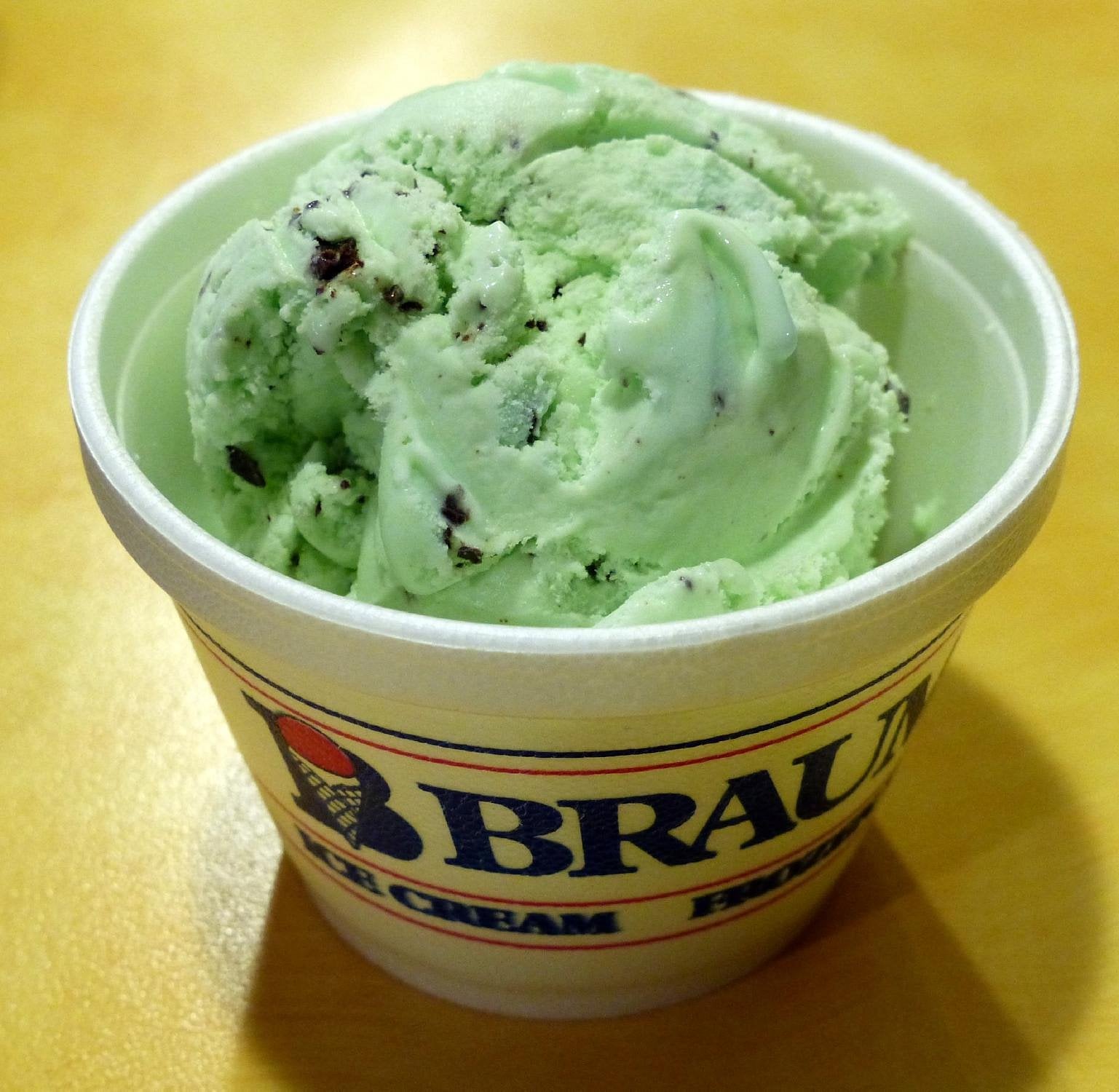 A white cup of mint chocolate chip ice cream, reminiscent of the sweet treats found at regional fast-food chains, sits on a light brown surface. The ice cream is pale green with small dark chocolate pieces, and the cup has a partial brand logo visible.