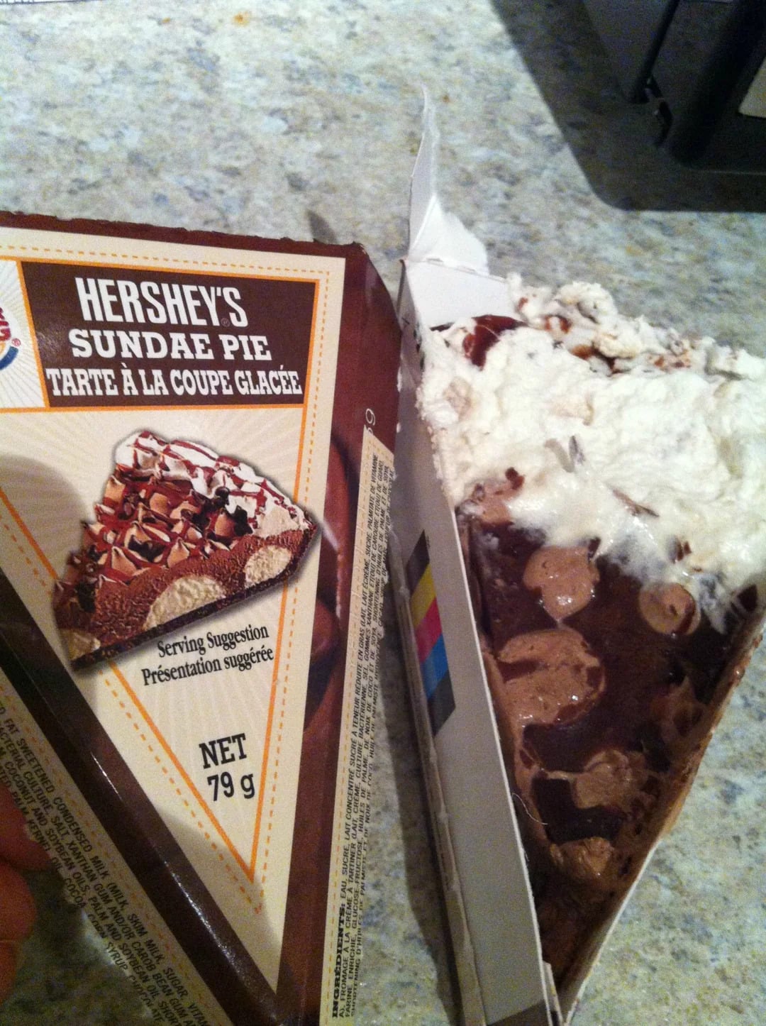A slice of Hershey's Sundae Pie in an open cardboard package sits on a countertop. The pie has a chocolate and cream topping with chocolate pieces, and the packaging displays an image of the pie and its nutritional information.