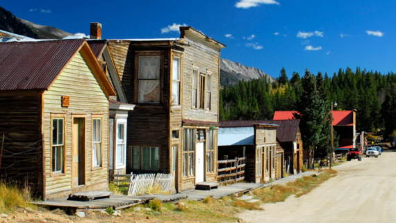 A row of old, weathered wooden buildings lines a dusty street in a small, historic western town&mdash;one of many classic American towns&mdash;set against mountains and pine trees in the background under a clear blue sky.