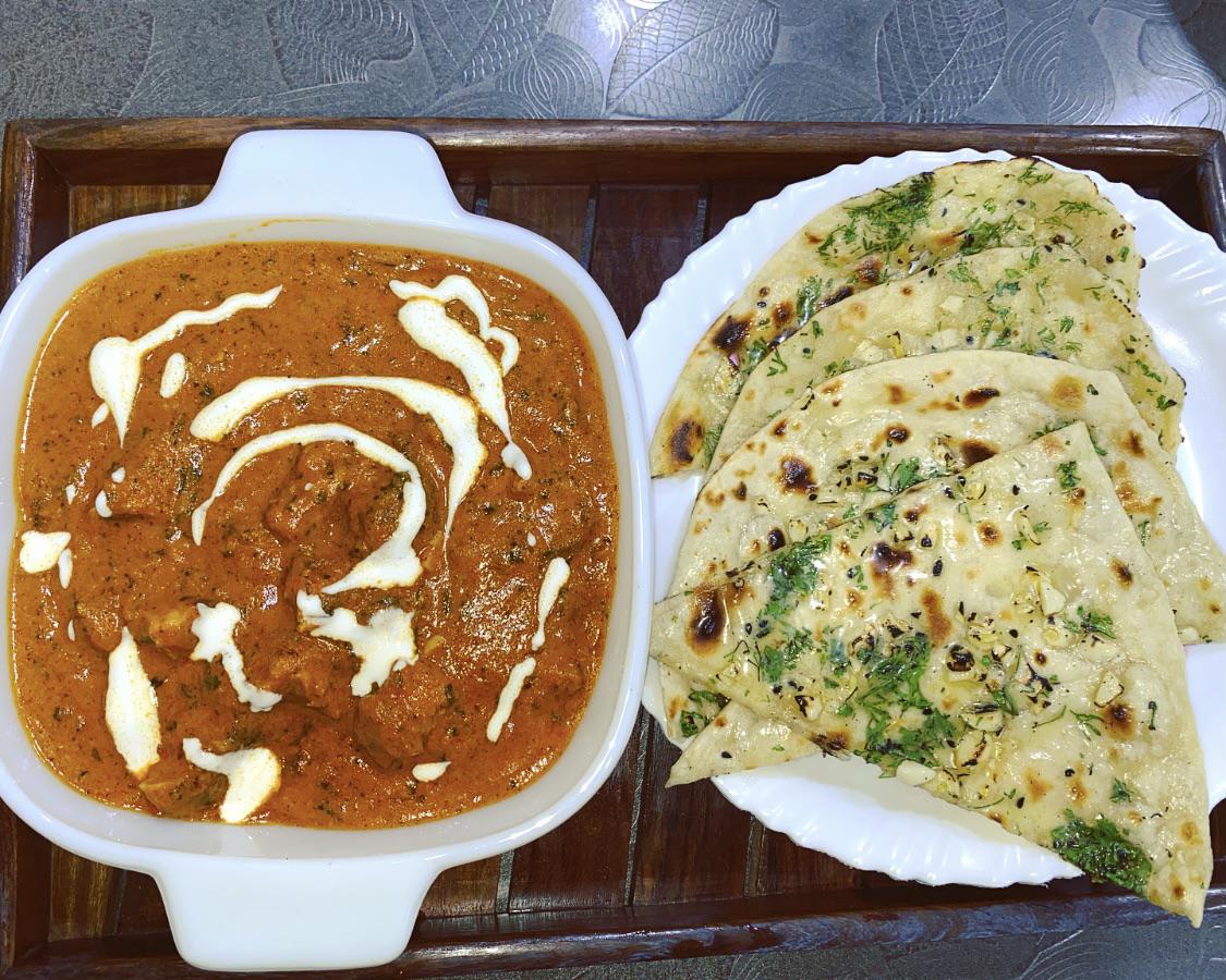 A bowl of creamy tomato-based curry garnished with cream, served with slices of garlic naan on a white plate, all placed on a wooden tray.