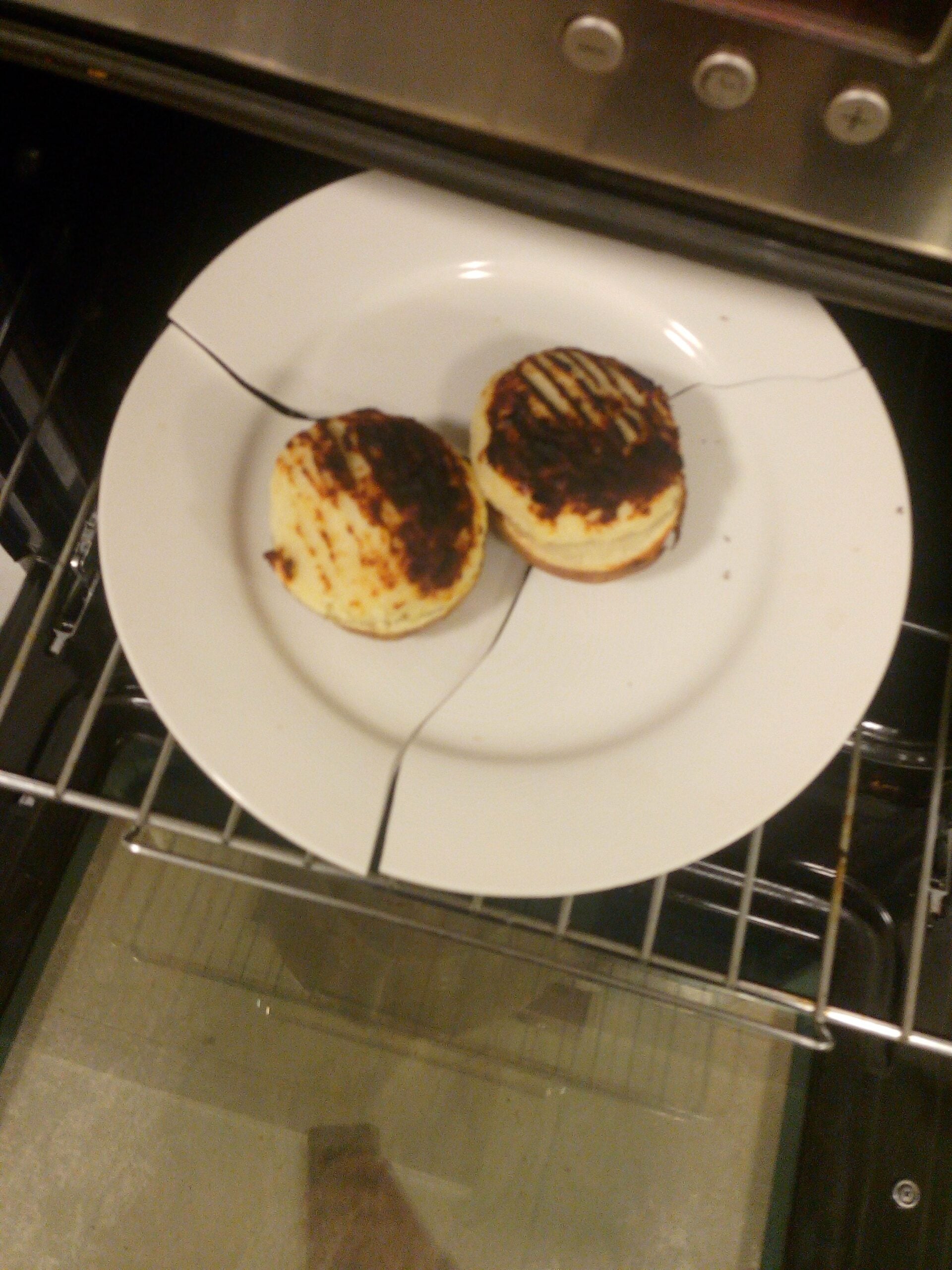 A cracked white plate with two burnt muffins sits on an oven rack, partially inside the oven.