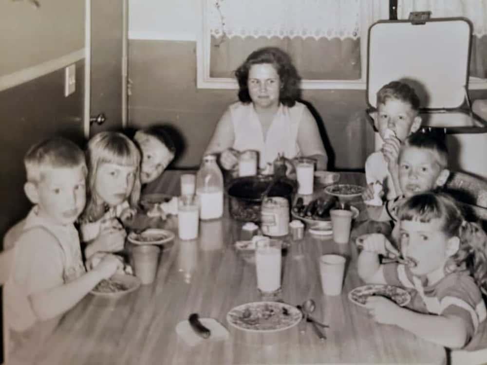 Six children sit around a table eating, while an adult woman sits at the head of the table. They appear to be having a meal together in a kitchen or dining area—a scene that captures the essence of nostalgic dinners. The photo is in black and white.