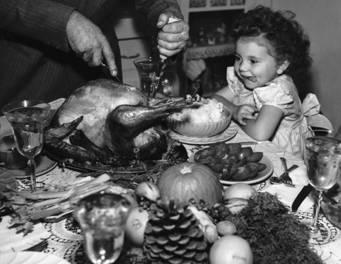 Black-and-white vintage photo of a man carving a roasted turkey at a table. A young, curly-haired girl sits nearby, watching with an excited, hungry expression as her tongue peeks out. The table is filled with festive food and decorations, capturing the warmth and joy of nostalgic dinners shared with loved ones.