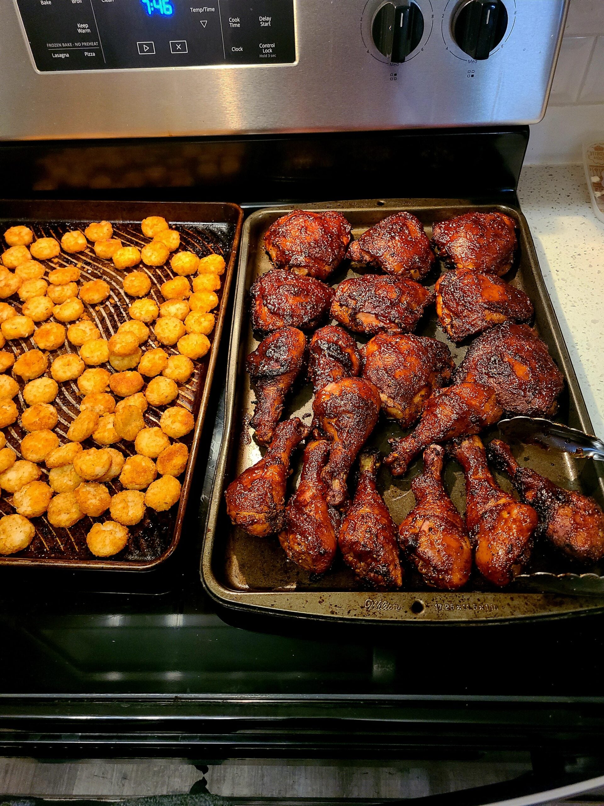A tray of crispy tater tots sits next to a pan filled with well-seasoned, baked chicken drumsticks and thighs on a stovetop.