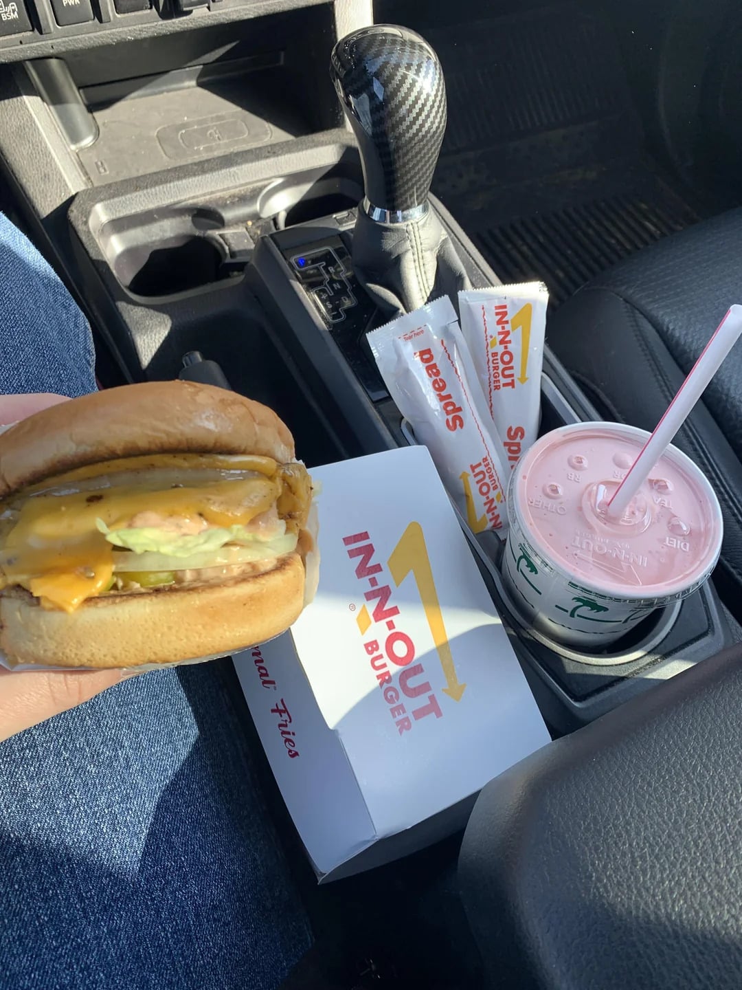 A hand holds a cheeseburger inside a car. Next to it are an In-N-Out box, a pink drink with straw, and three packets of Spread sauce on the car’s center console. Sunlight shines on the scene, highlighting these classic fast-food grocery store products for the perfect drive-through meal moment.