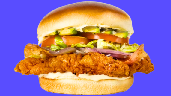 A crispy fried chicken sandwich with lettuce, tomato, pickles, onions, and bacon, topped with mayonnaise and served in a soft bun&mdash;just like the kind you&rsquo;d find at popular fried chicken chains&mdash;all set against a solid blue background.
