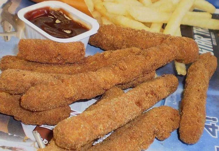 Breaded and fried mozzarella sticks are arranged on a tray with a container of marinara dipping sauce and a few French fries in the background.