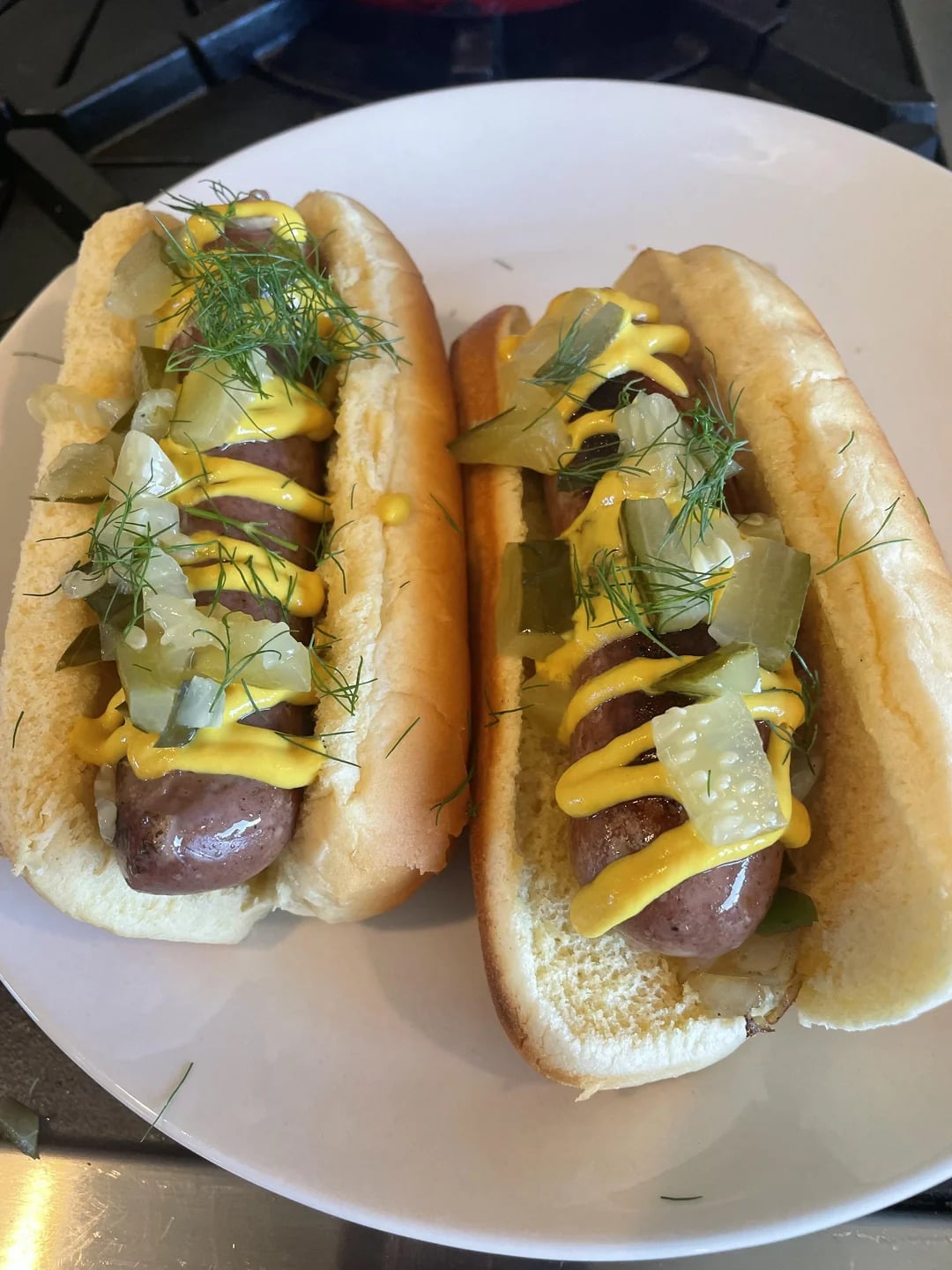 Two sausages in buns topped with diced pickles, yellow mustard, and fresh dill, served on a white plate.
