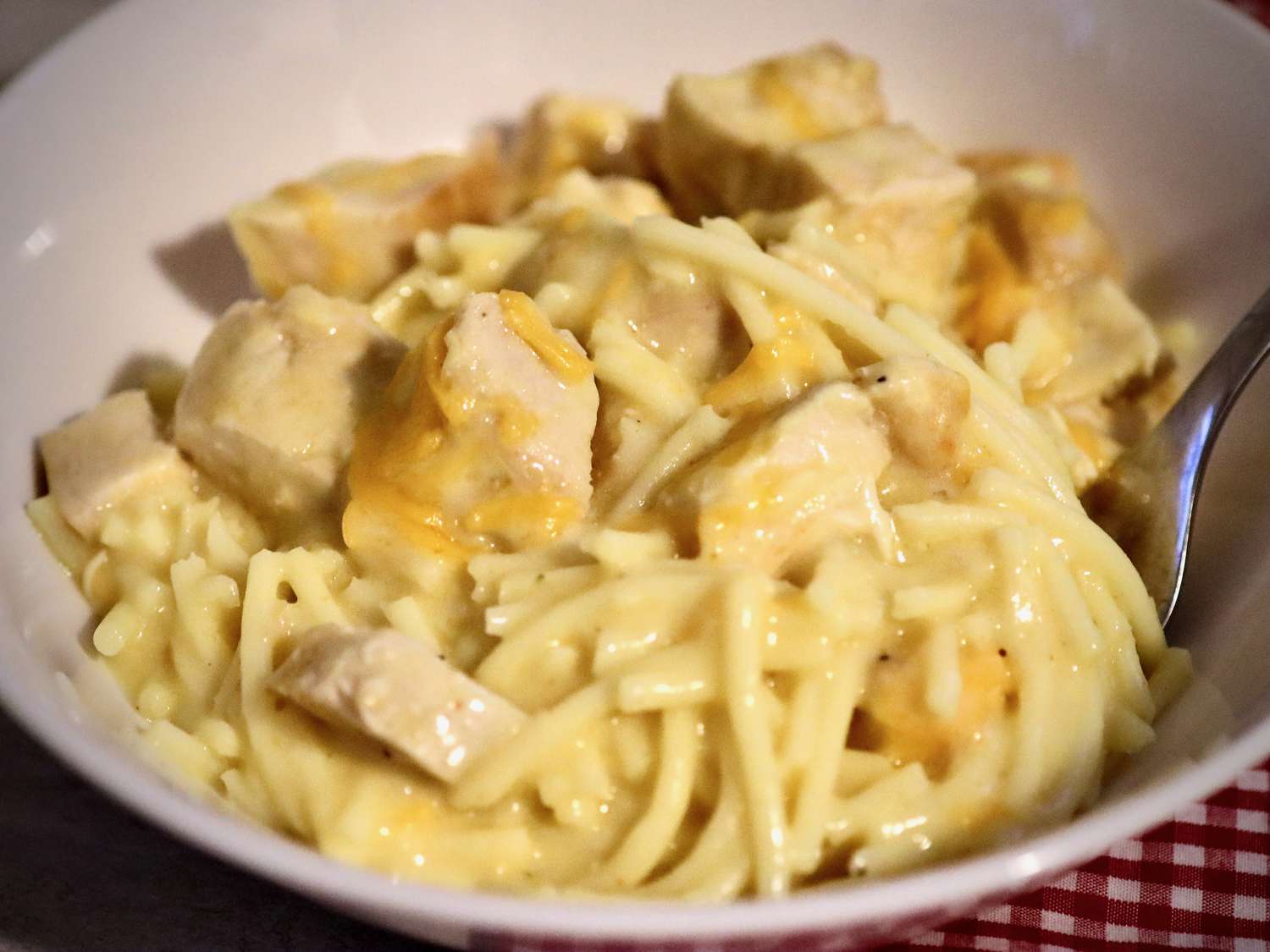 A bowl of creamy pasta with chunks of chicken, featuring spaghetti noodles coated in a rich cheese sauce. A metal fork is partially visible in the bowl.