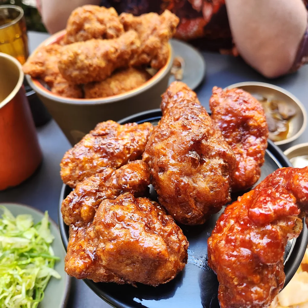 Close-up of crispy fried chicken wings, some glazed with sauce, served on a black plate. A bowl of more fried chicken, chopped greens, and small side dishes are in the background.
