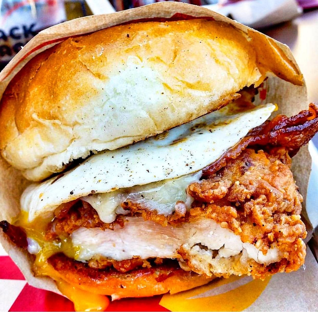 A close-up of a fried chicken sandwich with crispy bacon, a fried egg, melted cheese, and a soft toasted bun.