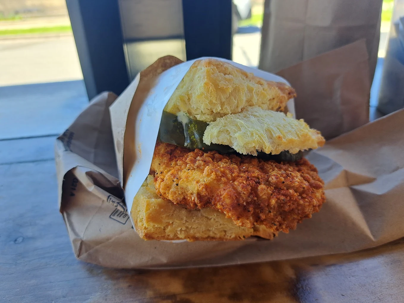 A close-up of a fried chicken sandwich with pickles, served on a flaky biscuit and wrapped in paper, sits on brown parchment paper on a wooden surface.