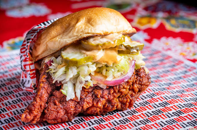 A spicy fried chicken sandwich with lettuce, pickles, onions, and sauce in a bun, wrapped partially in paper labeled "HOT!" on a red, decorated surface.