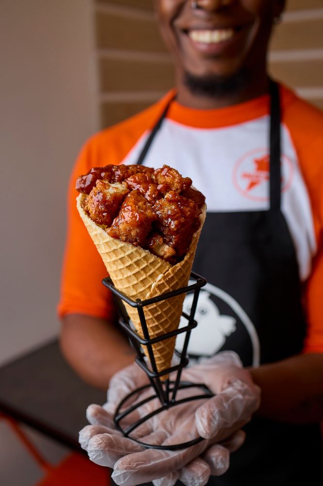 A smiling person in an apron holds a waffle cone filled with glazed fried chicken, presented in a black metal cone holder.