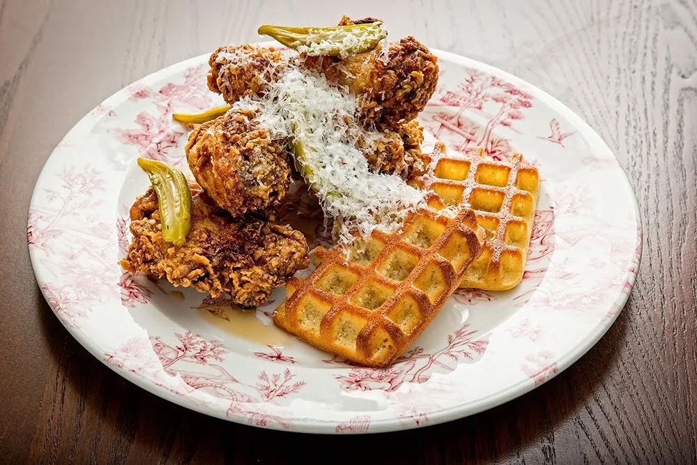 Fried chicken topped with shredded cheese and pickles, served with two pieces of golden brown waffles on a decorative plate, set on a wooden table.