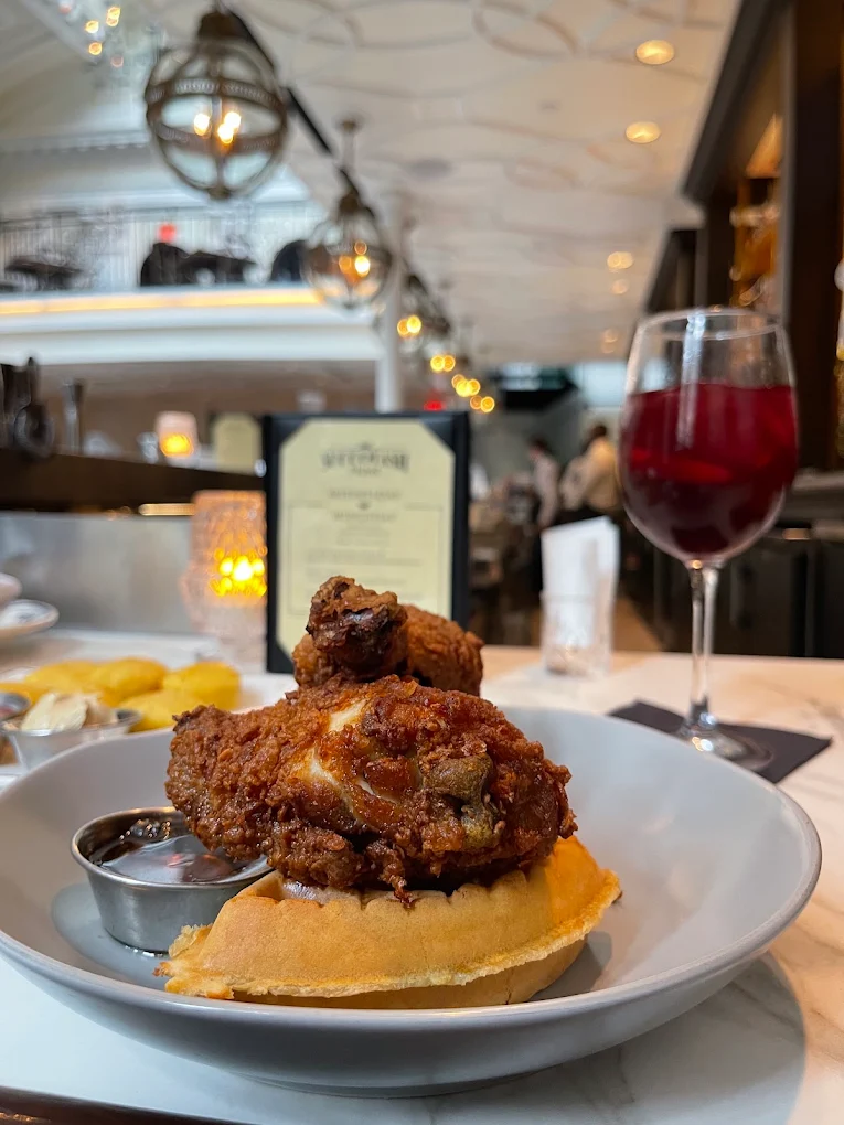 A plate of fried chicken on a waffle with sauce sits on a white table next to a glass of red drink in a stylish restaurant with warm lighting and modern decor.