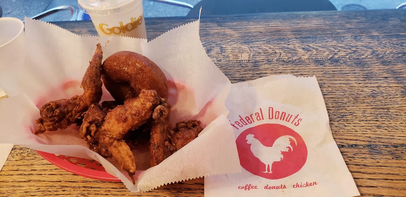 A basket with fried chicken wings and a donut on parchment paper, next to a Federal Donuts paper bag and a drink cup, all on a wooden table.