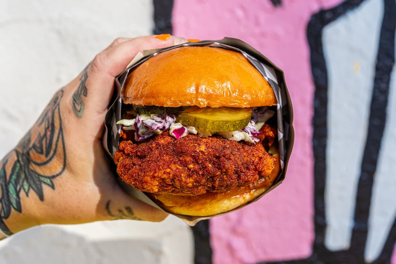 A tattooed hand holds a fried chicken sandwich with coleslaw, pickles, and a shiny brioche bun, in front of a colorful wall.