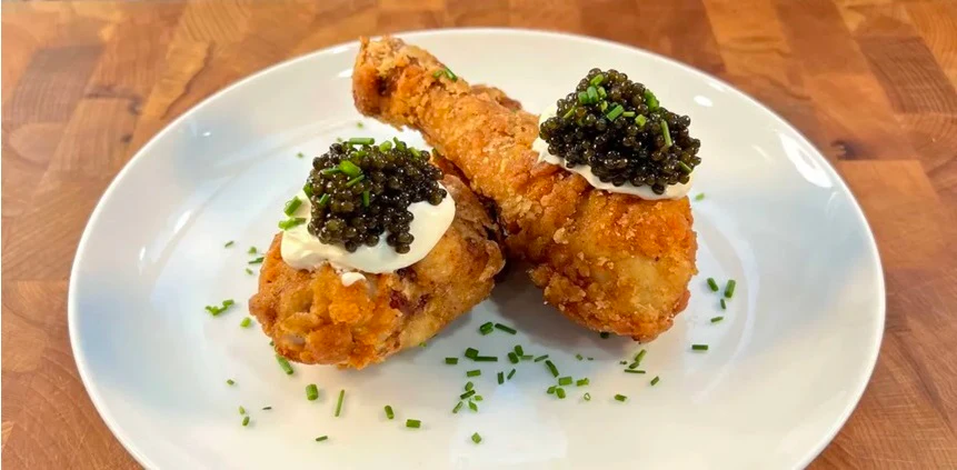 Two pieces of fried chicken on a white plate, each topped with a dollop of sour cream, black caviar, and chopped chives, sitting on a wooden surface.
