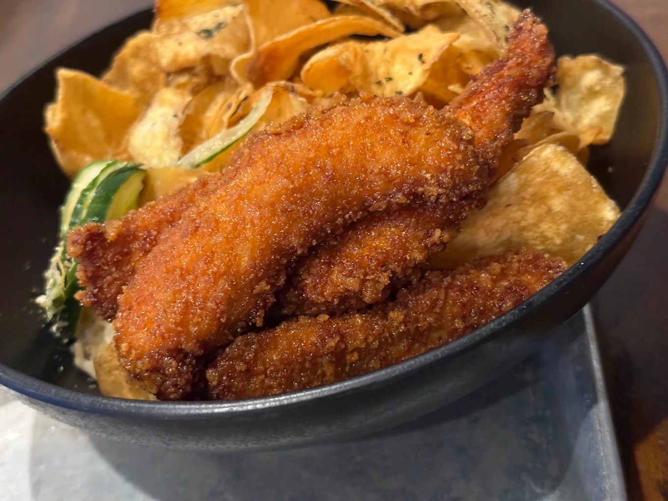A black bowl filled with crispy breaded chicken tenders, golden potato chips, and a few cucumber slices, served on a metal tray.