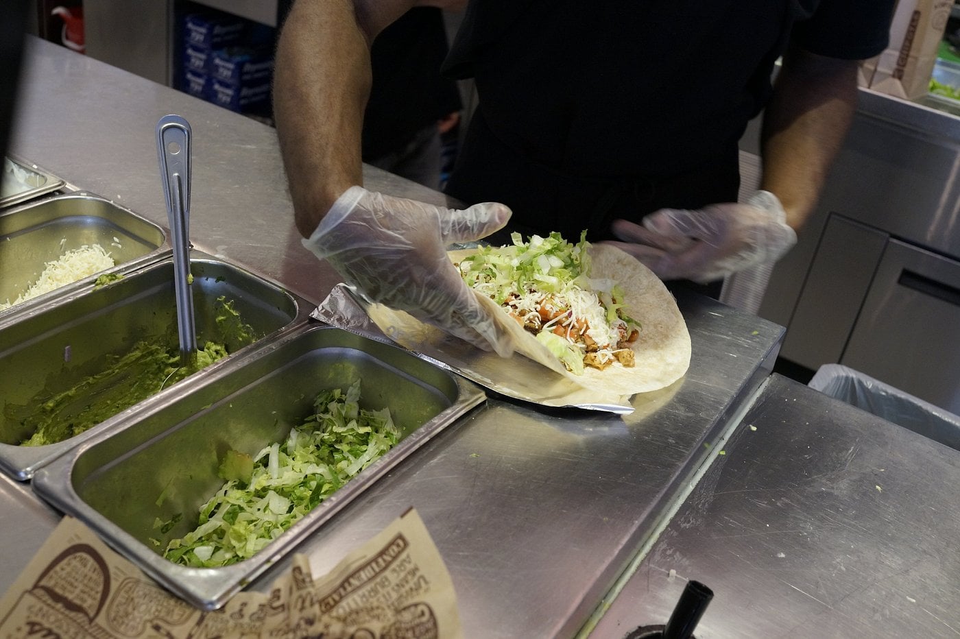 A person wearing gloves prepares a burrito with lettuce, cheese, and other ingredients at a restaurant counter, near containers of guacamole and shredded lettuce.