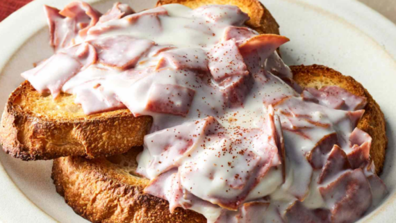 Two slices of toasted bread topped with chopped corned beef and creamy white sauce, lightly sprinkled with black pepper, served on a white plate&mdash;a classic reminiscent of hearty 1950s family meals.
