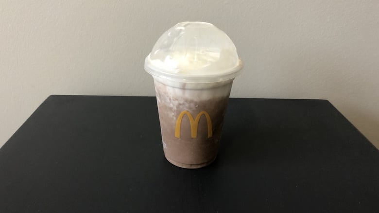 A McDonald's chocolate milkshake in a clear plastic cup with a dome lid and topped with whipped cream, sitting on a dark surface against a plain light-colored wall.