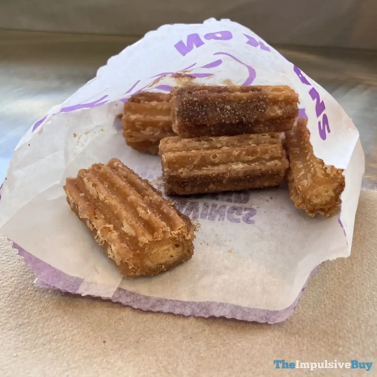 Five short churros with ridged sides, coated in cinnamon sugar, sit on a napkin and a partially opened white paper bag with purple text.
