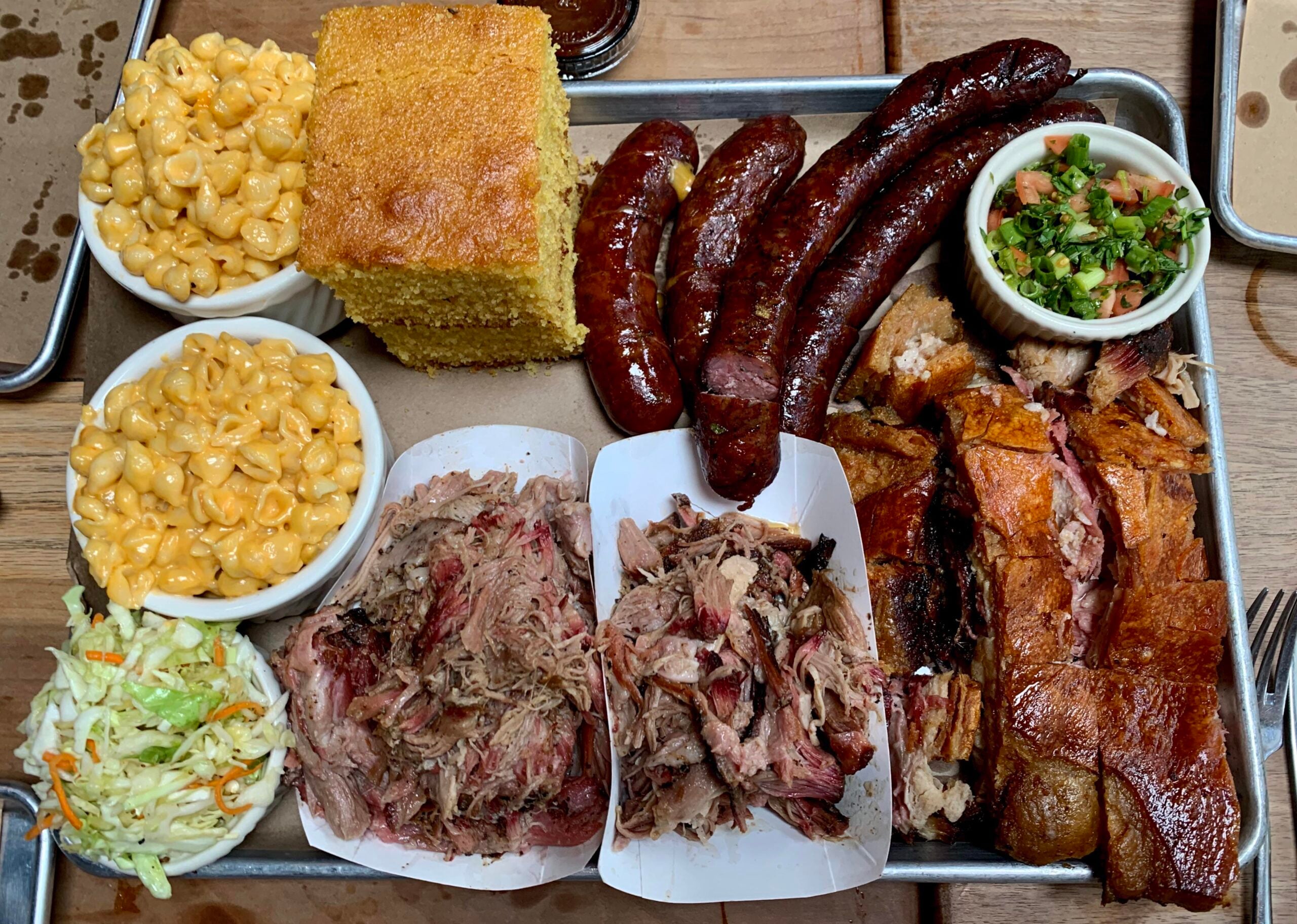 A tray with sliced brisket, sausages, pulled pork, mac and cheese, cornbread, coleslaw, roasted pork with crispy skin, and a small cup of salsa on a wooden table—everything you need for one of the best BBQ cookouts this summer.