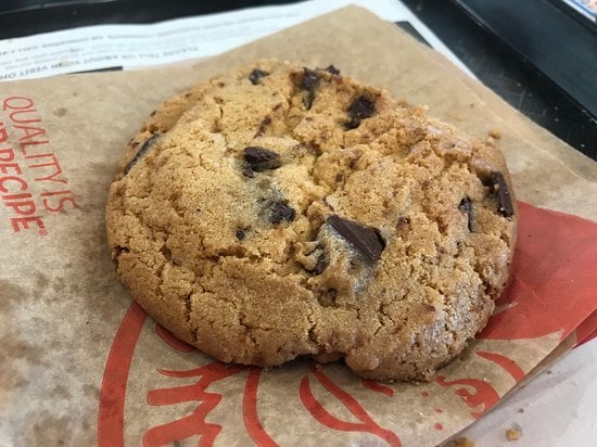 A large chocolate chip cookie with visible chocolate chunks sits on a brown paper wrapper with red print. The cookie appears golden-brown and freshly baked.