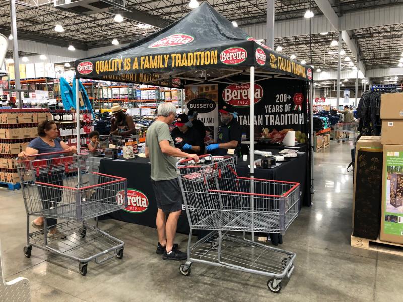 Shoppers sample food at a Beretta deli booth inside a warehouse store. Several people stand with shopping carts near the booth under a branded canopy promoting Italian tradition.