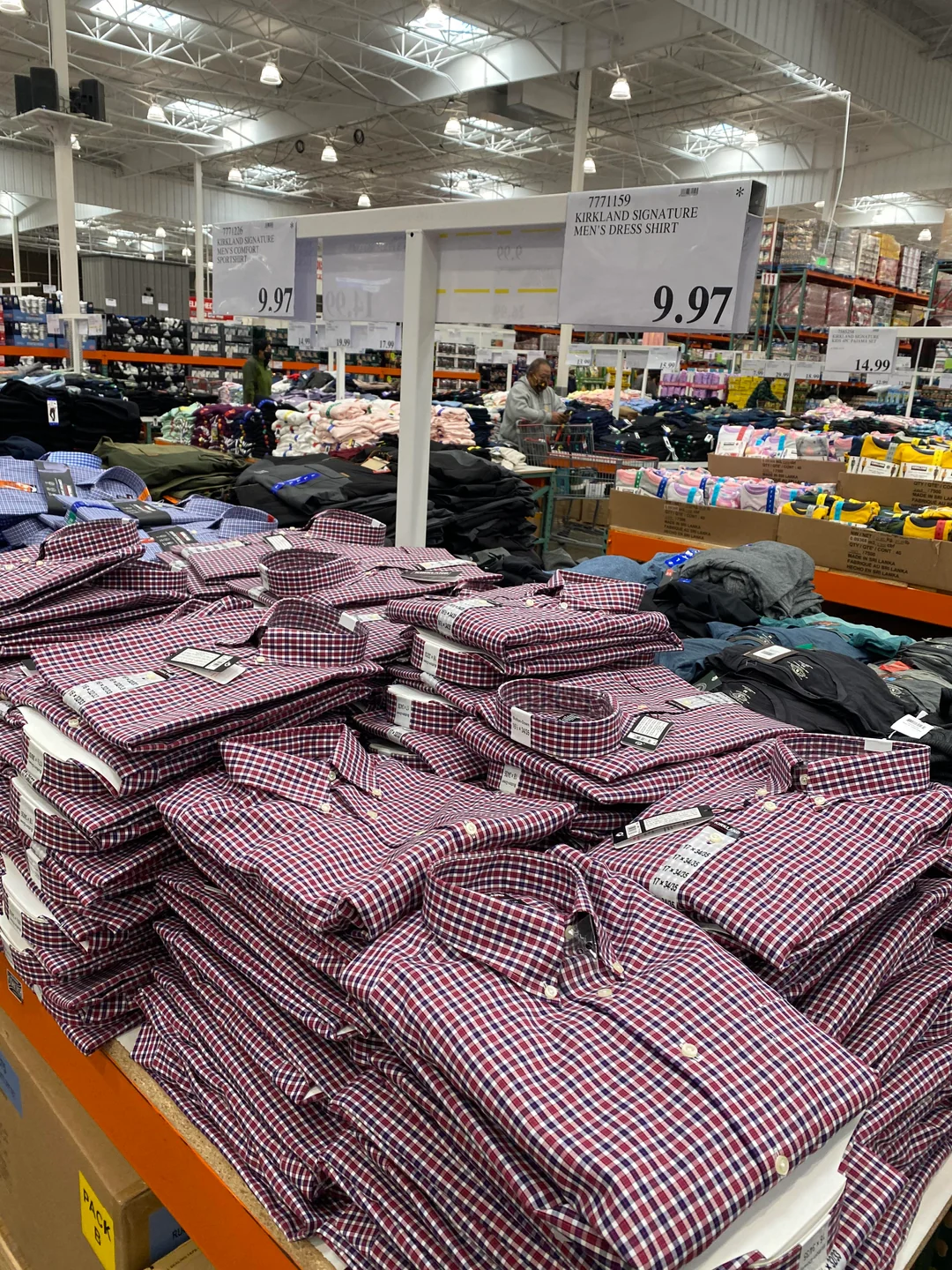 Stacks of folded men's plaid dress shirts on a display table in a warehouse store, with more clothing, products, and shoppers visible in the background. A sign lists the price as $9.97.