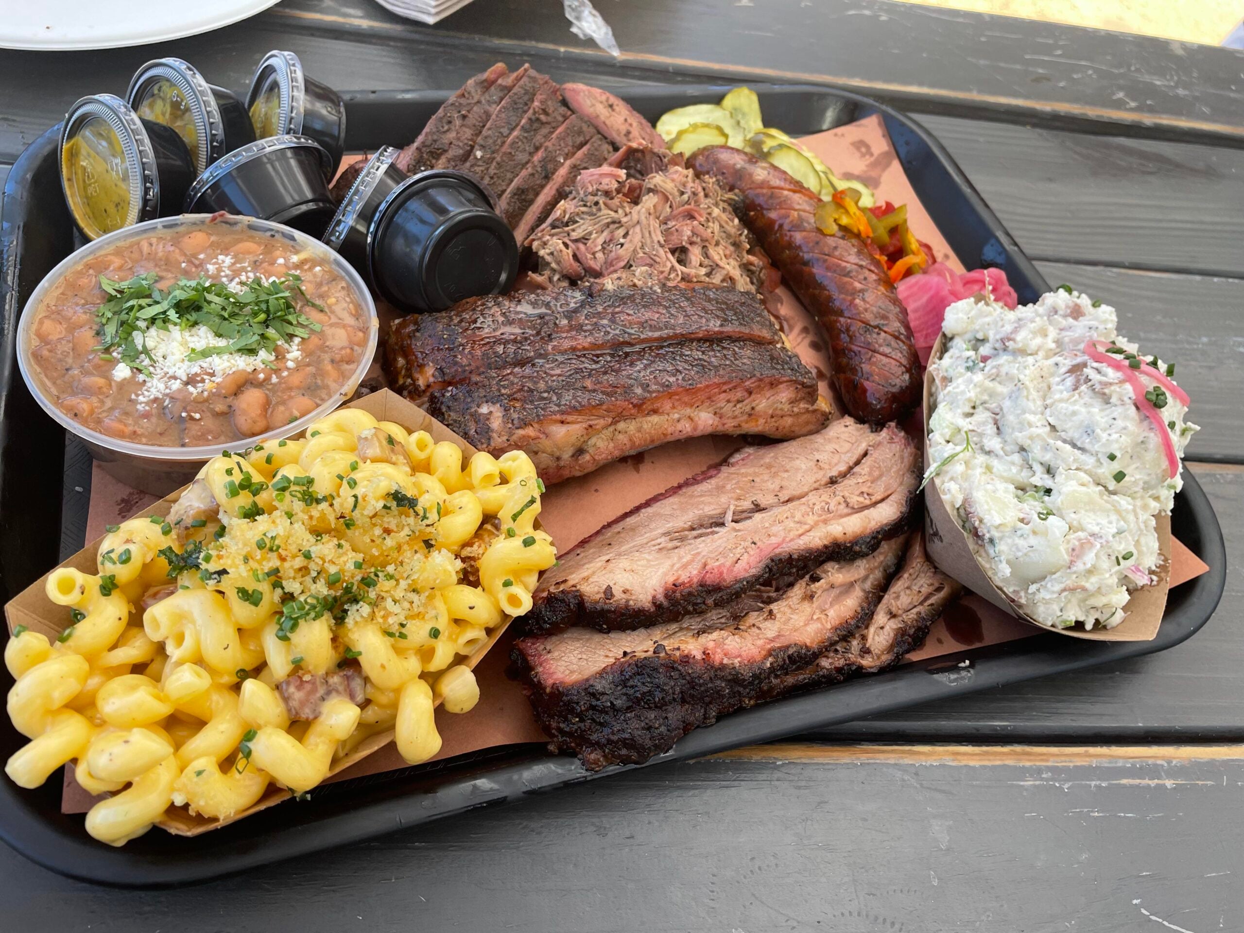 A tray of barbecue food with brisket, ribs, sausage, pulled pork, baked beans, macaroni and cheese, potato salad, pickles, and sauce cups on a black picnic table—perfect for the best BBQ cookouts this summer.