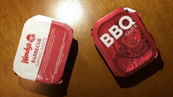 Two square packets of barbecue dipping sauce are on a wooden surface. The left packet is labeled "Wendy's Barbecue Dipping Sauce," and the right packet is labeled "BBQ Sauce" with a red design.
