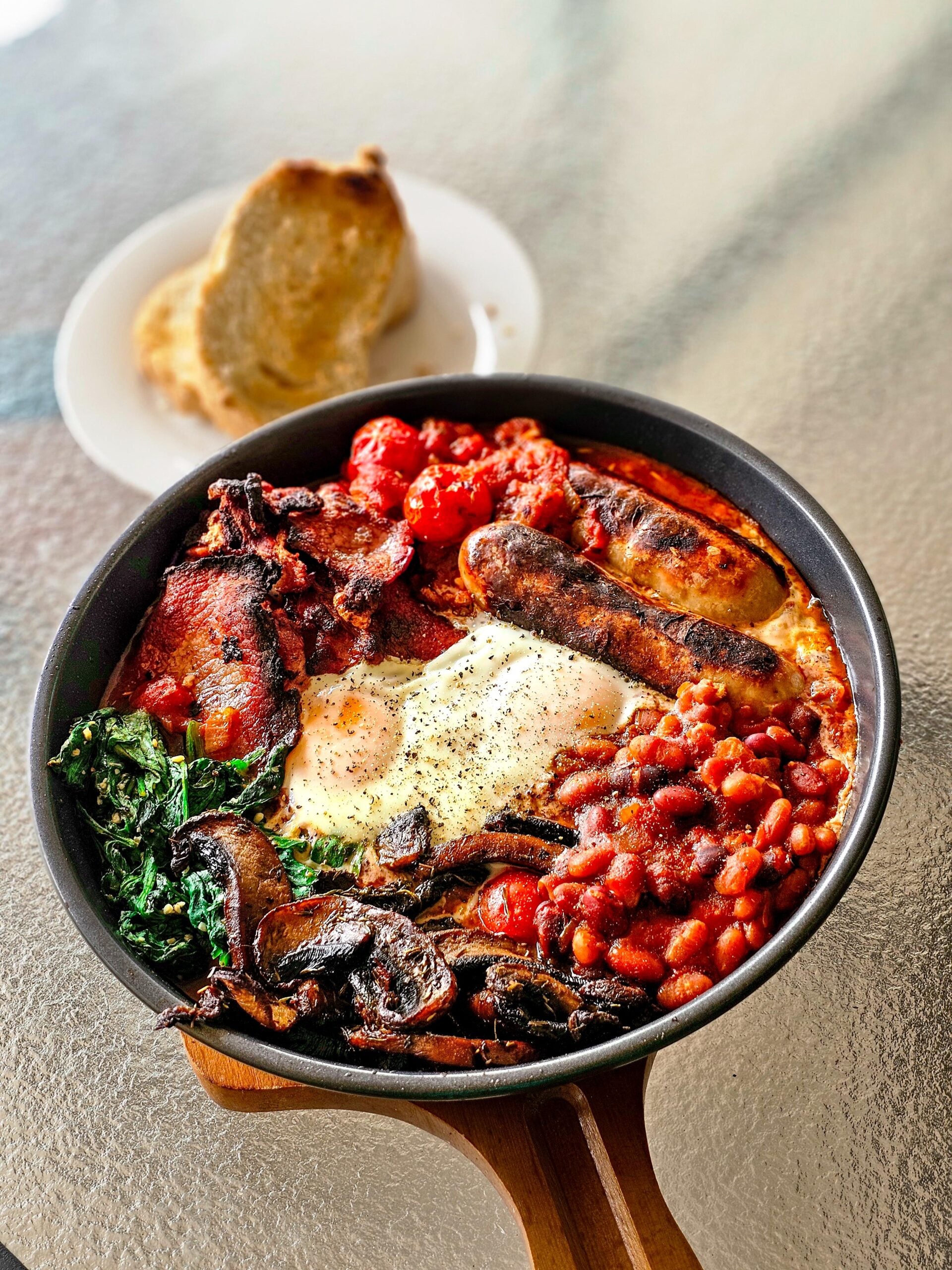 A skillet filled with fried eggs, sausages, bacon, beans, tomatoes, spinach, and mushrooms sits on a table, with a plate of bread slices in the background.