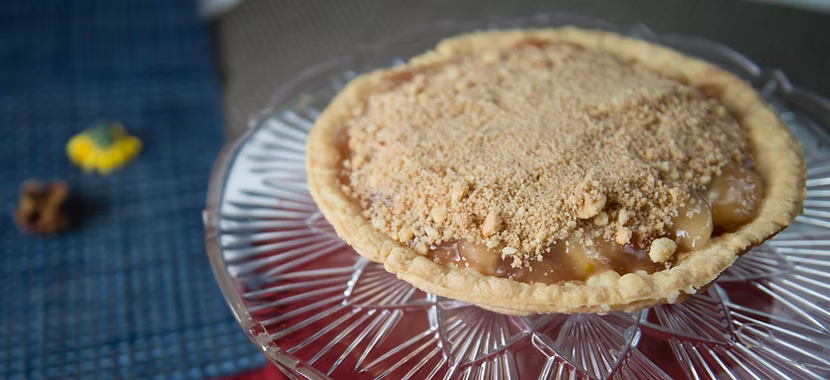A crumb-topped apple pie sits on a glass cake stand, displaying the classic golden crust and generous crumbly topping that make apple pies irresistible. The pie filling peeks out beneath, with a blue placemat and small blurred objects in the background.