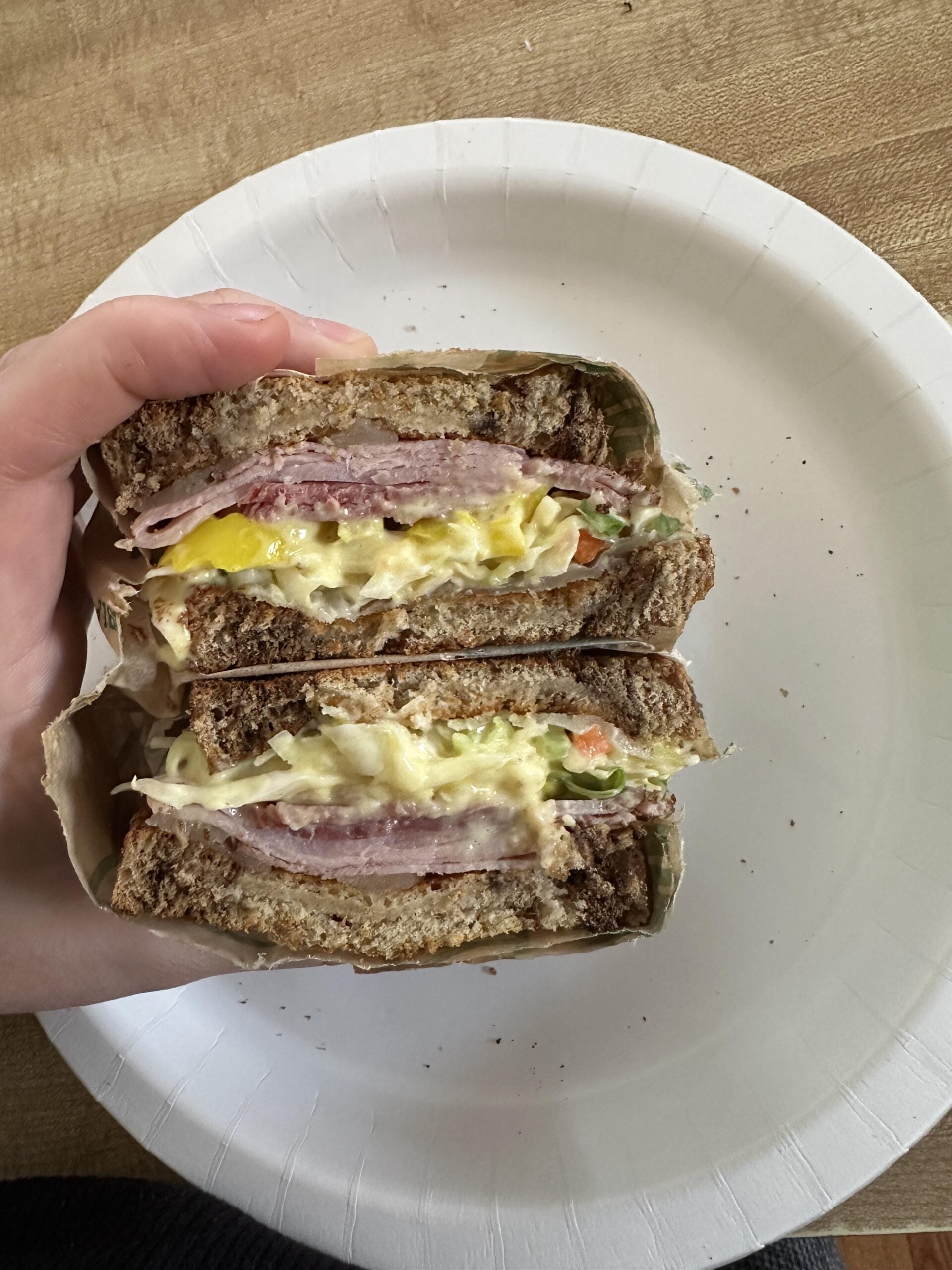 A hand holds a sandwich cut in half, showing layers of ham, egg, lettuce, tomato, and cheese on brown bread. The sandwich rests on a white paper plate on a wooden surface.