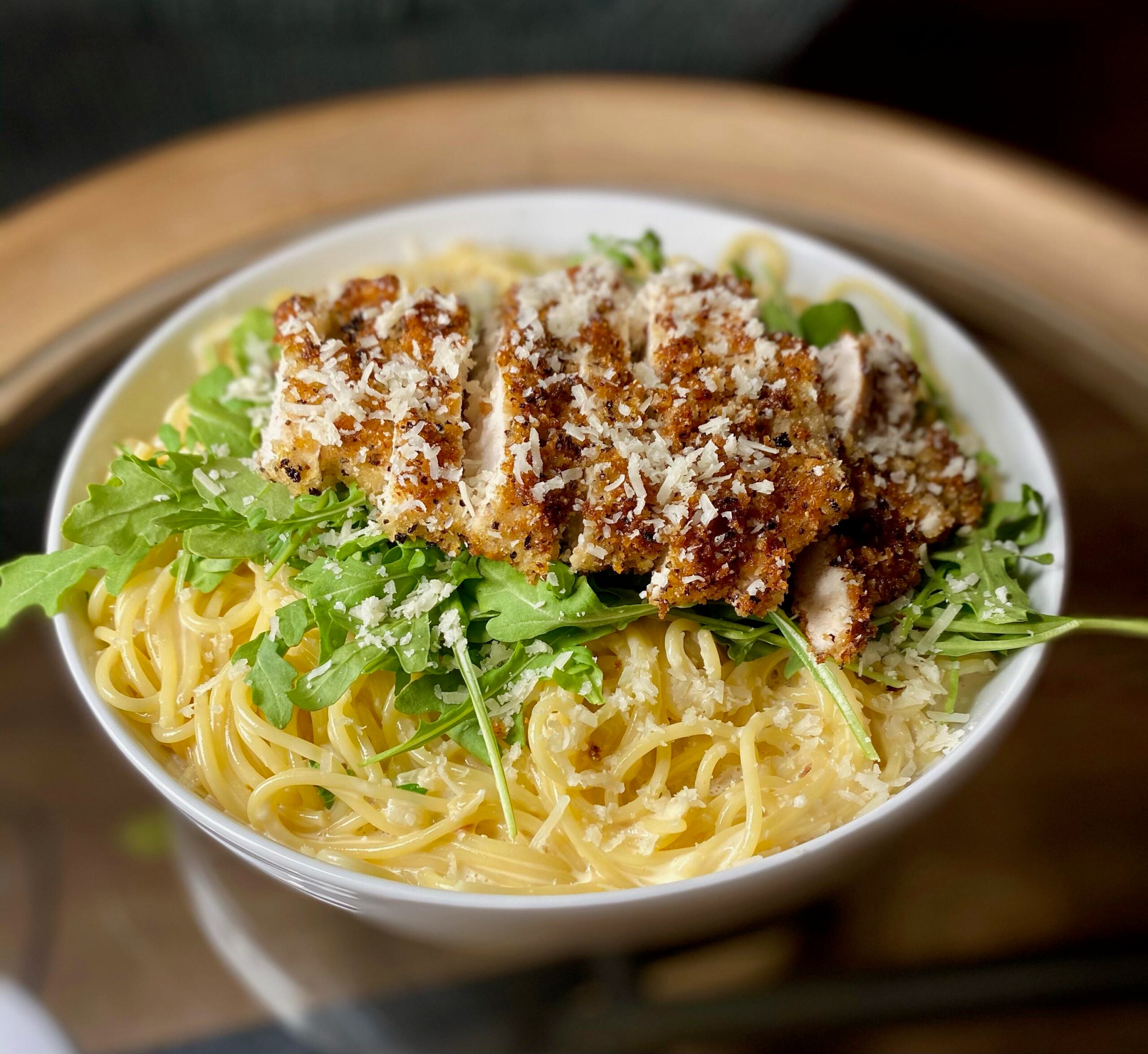 A bowl of spaghetti topped with crispy breaded chicken slices, fresh arugula, and grated parmesan cheese, served on a wooden tray.