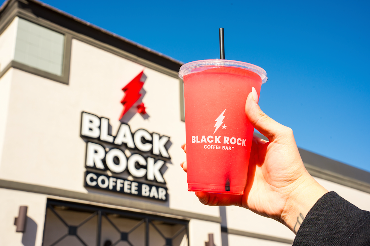 A hand holds a red drink in a clear cup with a black straw, displaying the Black Rock Coffee Bar logo, in front of one of the brand’s vibrant coffee houses on a sunny day.