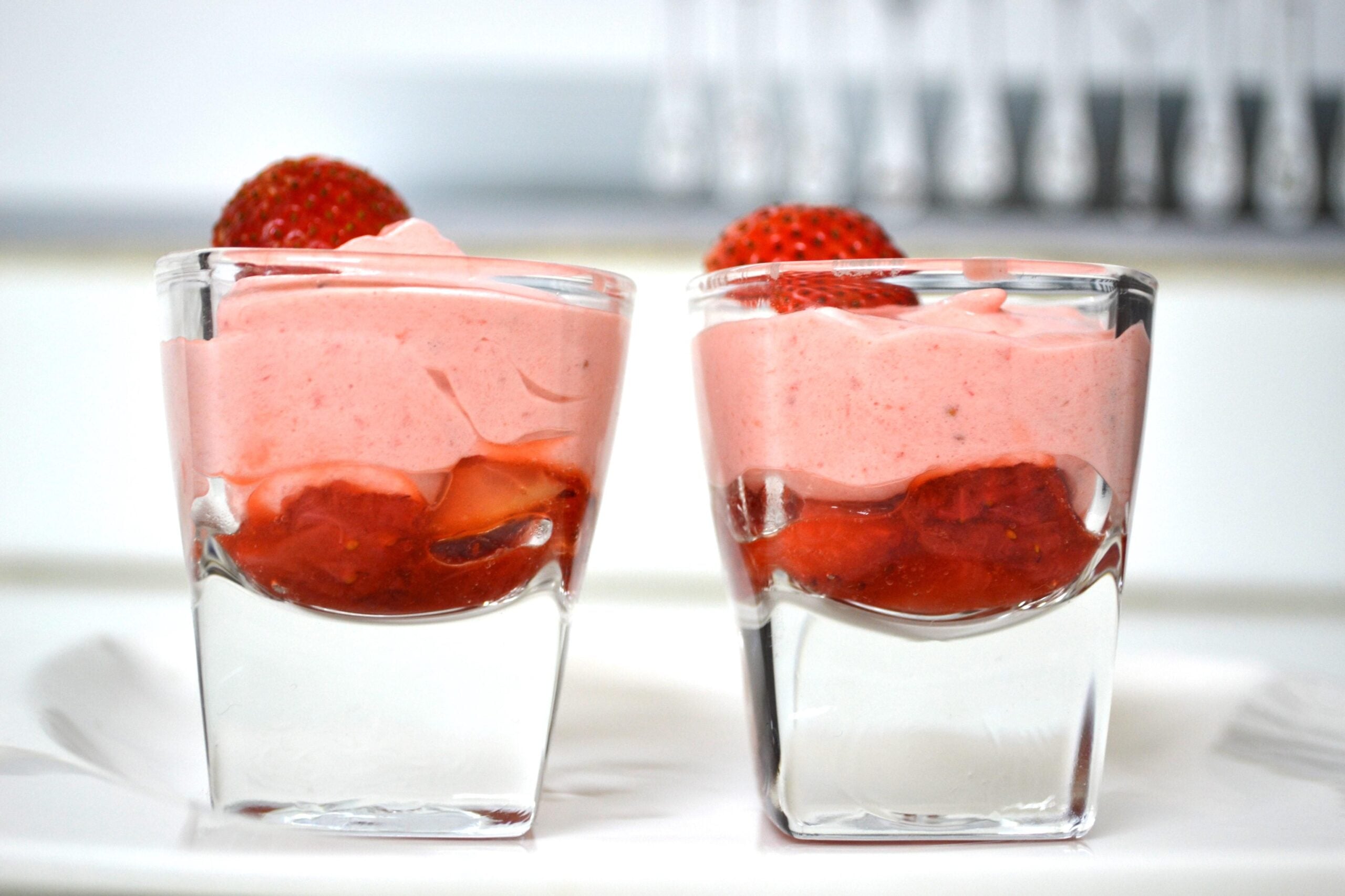 Two small glass cups filled with layers of pink strawberry mousse and strawberry pieces, each topped with a whole strawberry, showcase the charm of homemade desserts, set on a white surface with a blurred background.