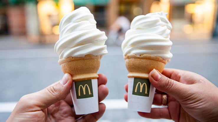 Two hands holding McDonald's soft serve ice cream cones with white swirl tops and classic wafer cones, against a blurred street background.