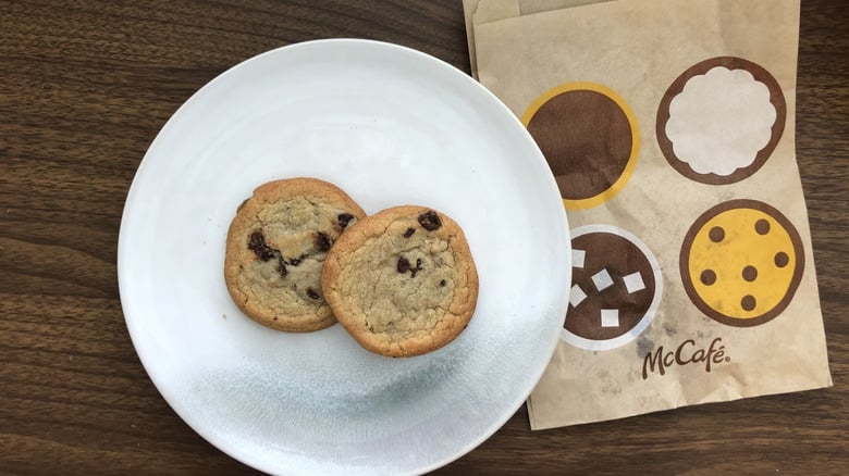 Two chocolate chip cookies on a white plate, next to a McCafé paper bag with illustrated cookies, sugar cubes, and a pie on a brown wooden table.